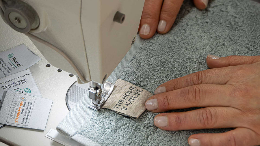 A seamstress at a sewing machine, sewing a THON label onto a TENCEL™ Lyocell and Cotton blue towel.