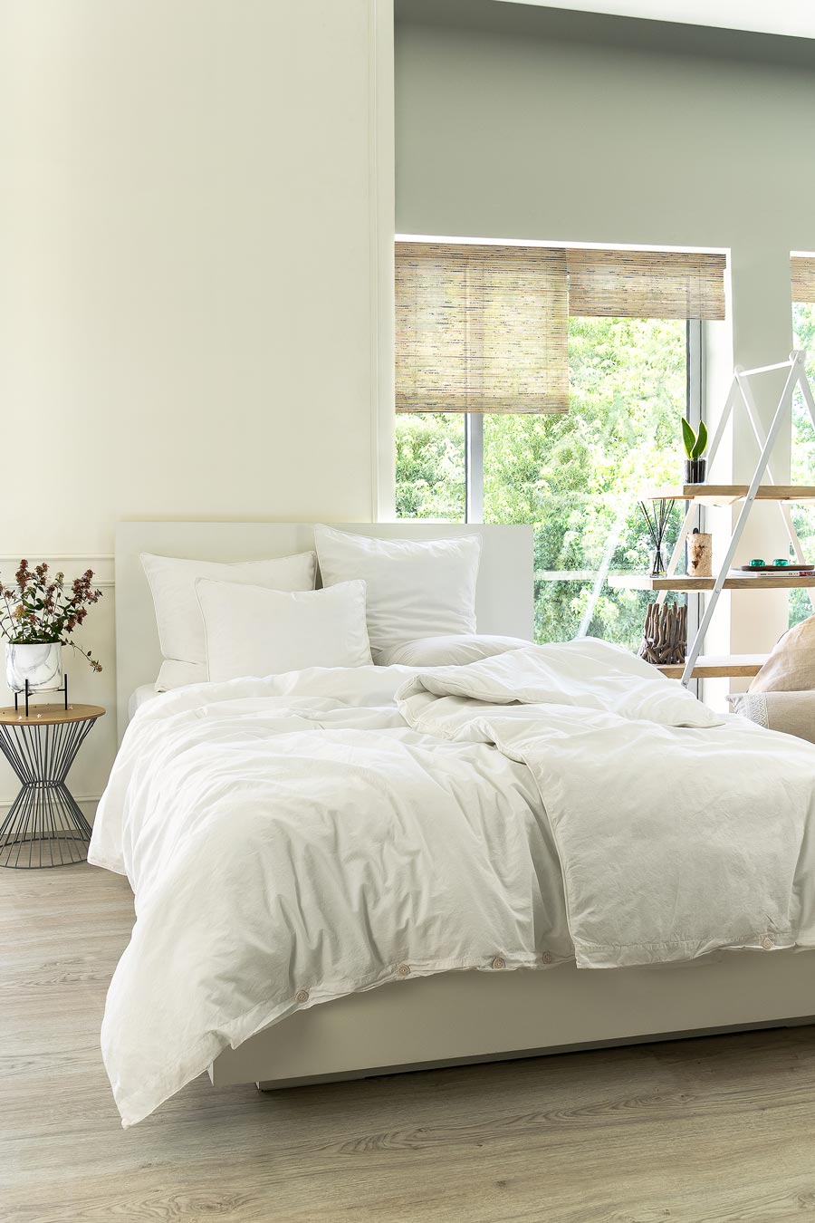 Bed with white Organic Cotton Percale bedding next to a shelf and bedside table with plants.